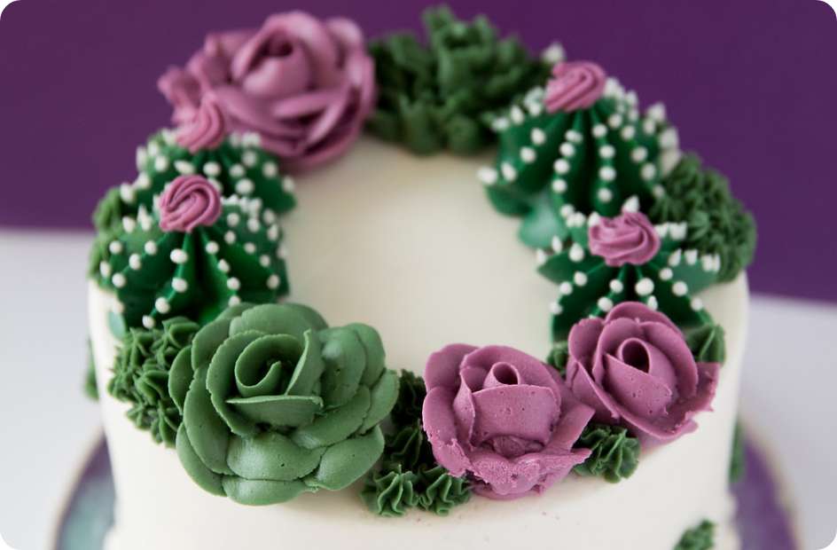 A vanilla cake iced with white buttercream. There are green  leaves piped around the top and sides and purple roses piped on the top around the perimeter of the cake